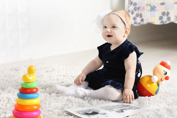 Little cute baby in dress sits on soft carpet among toys and smi — Stock Photo, Image