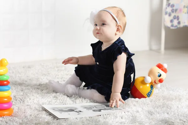 Little cute baby in dress sits on soft carpet among toys and loo — Stock Photo, Image