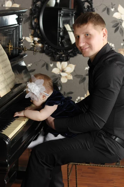 Smiling man in black with cute baby sits at piano in room. Baby — Stock Photo, Image