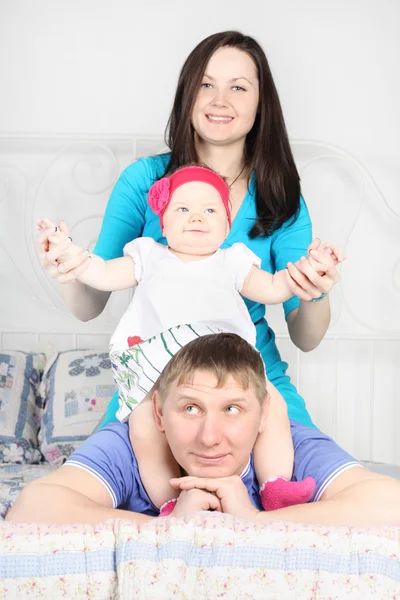 Beautiful mother and happy baby sit on father lying on bed at ho — Stock Photo, Image