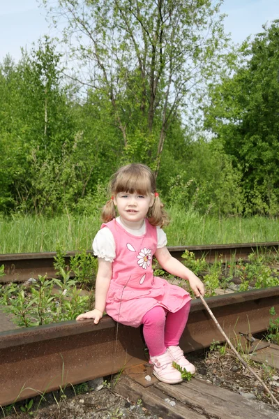 Little cute girl in pink dress with stick sits on old railway at — Stock Photo, Image