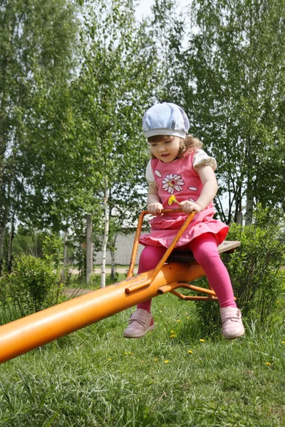 Happy little beautiful girl in pink rides on orange seesaw at su — Stock Photo, Image