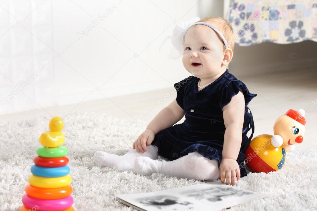 Little cute baby in dress sits on soft carpet among toys and smi
