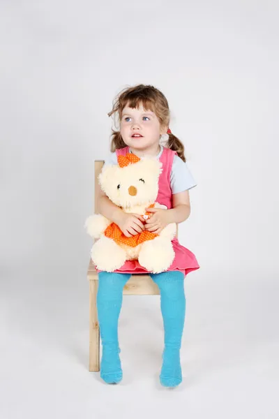 Little cute girl sits at wooden chair with toy bear and looks aw — Stock Photo, Image