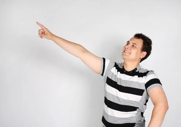 Brunet man in striped T-shirt smiles and points to side on grey — Stock Photo, Image