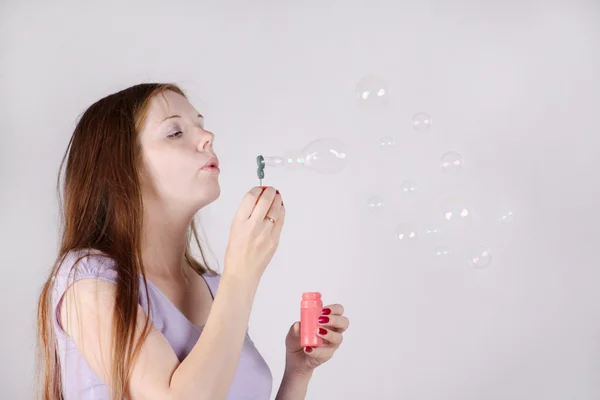 Beautiful woman with long hair blows soap bubbles on grey backgr — Stock Photo, Image