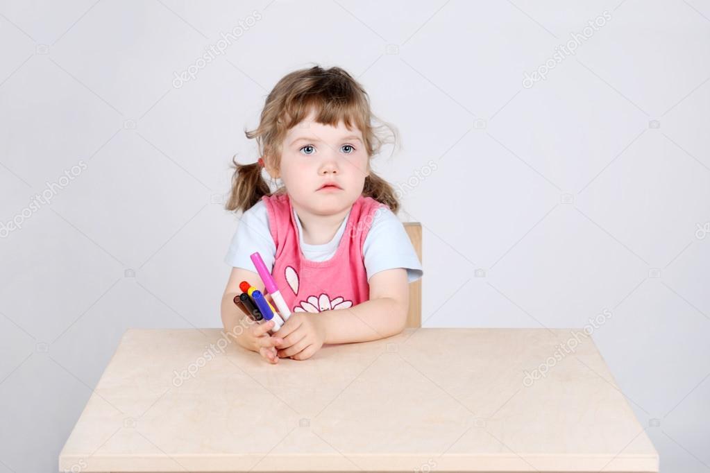 Little sad girl sits at wooden table and holds with felt-tip pen