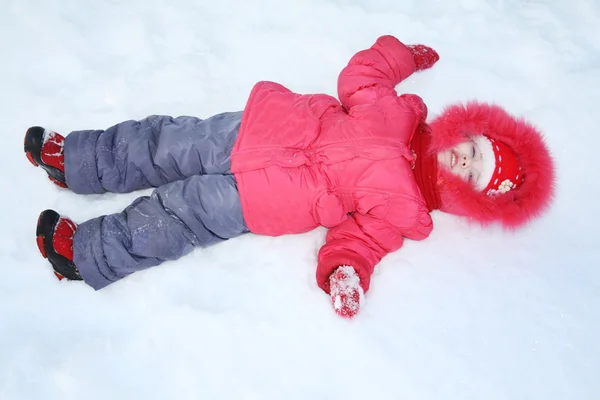 Glückliches kleines Mädchen in warmer Kleidung liegt im Schnee am Wintertag. — Stockfoto