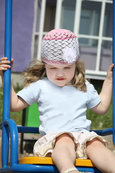 Pequena menina feliz senta-se no balanço no parque infantil edifício violeta em — Fotografia de Stock