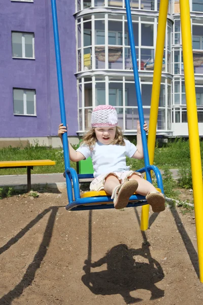 Little cute girl sits on swing at playground near building at su — Stock Photo, Image