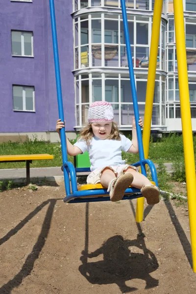 Niña linda se sienta en el columpio en el patio de recreo cerca del edificio en su — Foto de Stock