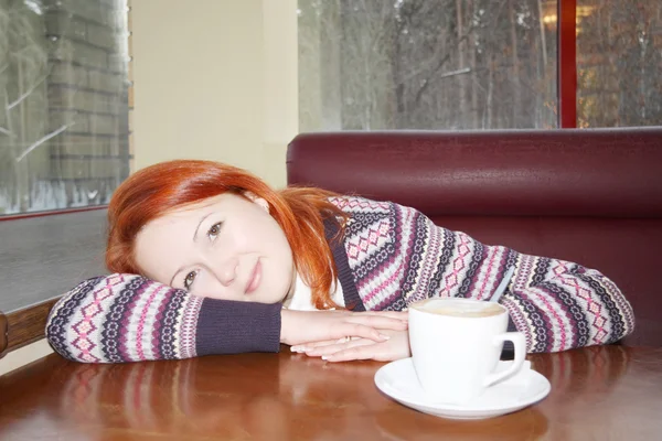 Zrzka šťastná dívka sedí u stolu v kavárně s bílým šálek cappuccino a vypadá to na kameru. — Stock fotografie