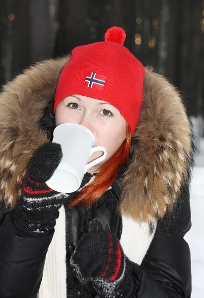 Happy girl in red hat drinks hot tea and looks at camera in fore — Stock Photo, Image