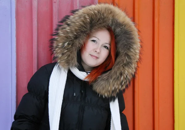 Girl in hood with fur stands near multicolored fence at winter. — Stock Photo, Image