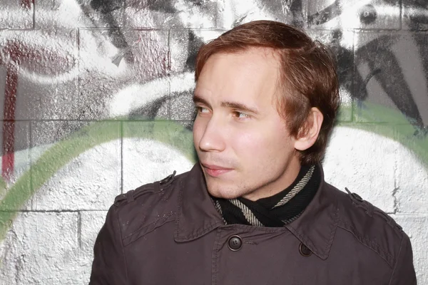 Jeune homme se tient près du mur avec des graffitis et regarde de côté en plein air — Photo