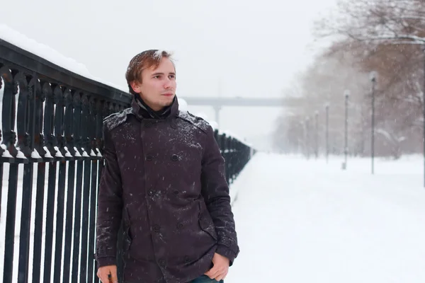 Young man with snowflakes in hair stands near metal black fence — Stock Photo, Image