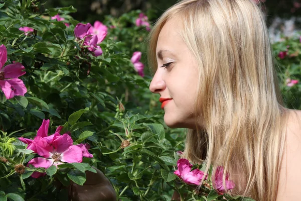 Beautiful blonde girl looks at pink flowers on bush in park at s — Stock Photo, Image