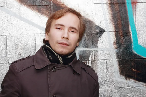 Young man stands near wall with graffiti and looks at camera out — Stock Photo, Image