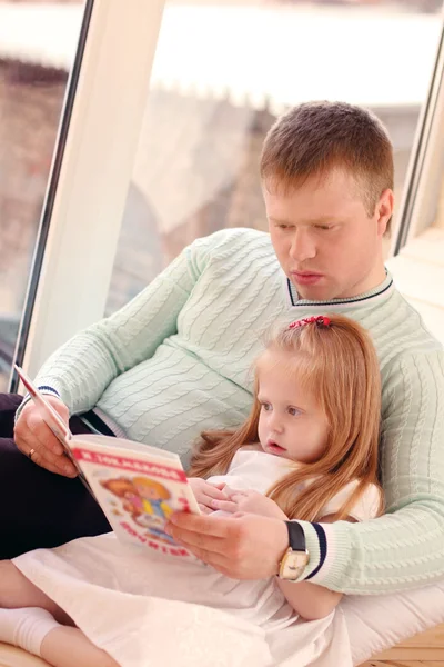 Pretty little blonde girl and father sit near window and read bo — Stock Photo, Image