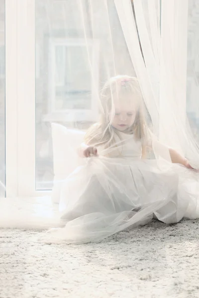 Pretty little blonde girl sits near big window and hides behind — Stock Photo, Image