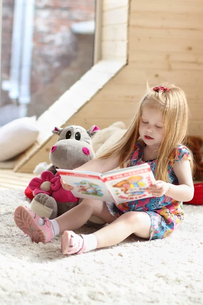 Pretty little blonde girl sits near soft toy on carpet in cozy r — Stock Photo, Image