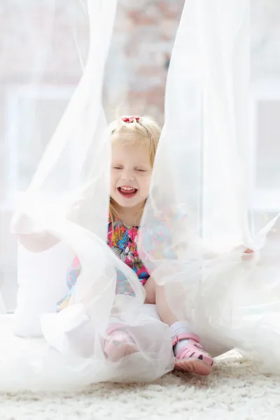 Hübsches kleines blondes Mädchen sitzt am großen Fenster, lacht und spielt — Stockfoto