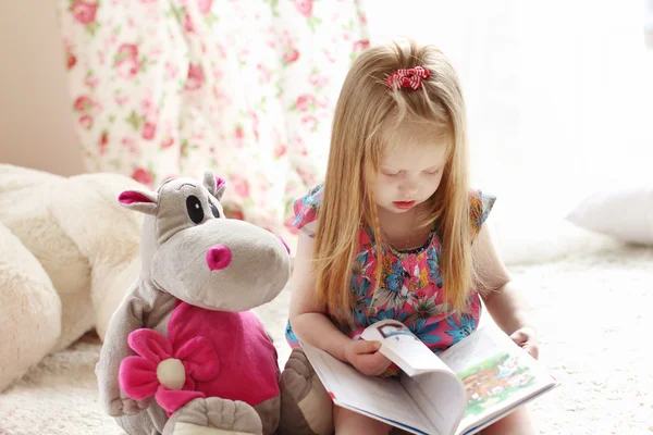 Bella bambina bionda siede vicino al giocattolo morbido sul tappeto e legge — Foto Stock