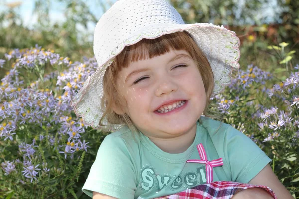 Little cute girl in white hat smiles near flowers at sunny day — Stock Photo, Image