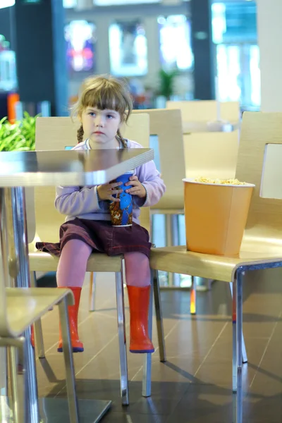 Little girl sitting at a cafe table with popcorn and soda — Stock Photo, Image