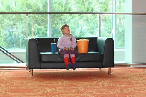 Little girl sitting on black sofa with popcorn and soda — Stock Photo, Image