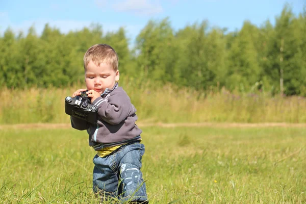 Little handsome boy holds camera and walks at meadow in summer d — Stock Photo, Image