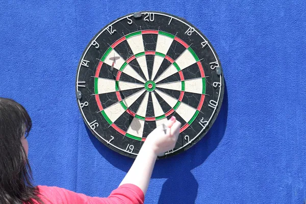 Woman removes darts from dartboard on blue wall at sunny day — Stock Photo, Image