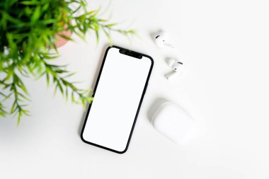 Beyaz masada beyaz ekran ve bluetooth kulaklıklı bir akıllı telefon var..