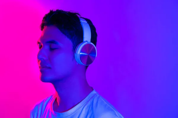 Music streaming services. Portraif of smiling handsome asian man in wireless headphones enjoying his favorite playlist, standing with closed eyes under neon lighting over purple background.