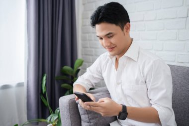 Yakışıklı Asyalı adam evde koltuğa yaslanırken akıllı telefon kullanıyor.
