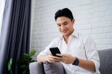 Mutlu bir genç Asyalı adam evde bir kanepede otururken cep telefonuyla portresi