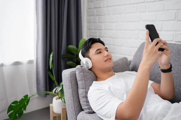 自宅のソファに横になっている間に スマートフォンを持ち ワイヤレスヘッドフォンを着用し 彼女の好きな音楽を聞いているカジュアルなTシャツを着た幸せな男30代の写真 — ストック写真