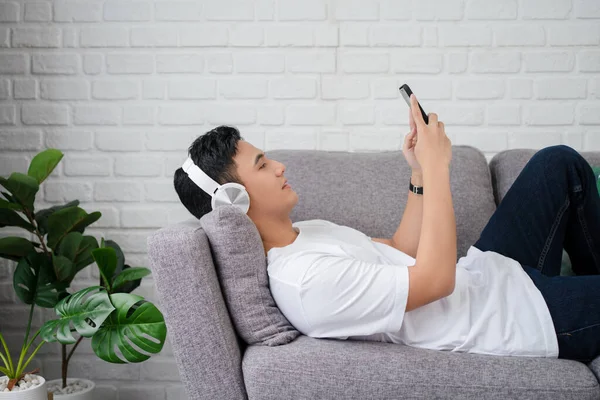 カジュアルなTシャツを着たハンサムな若い男 ソファに横になっている間 彼はスマートフォンを使って好きな音楽を聴いていた — ストック写真