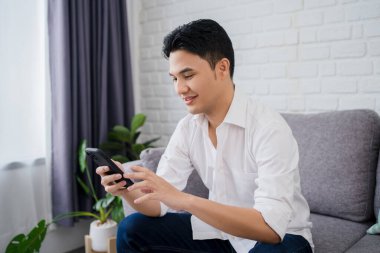 Mutlu gülümseyen genç erkek akıllı telefon kullanarak evdeki rahat koltuğun keyfini çıkarıyor.