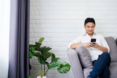Evdeki kanepede otururken gülümseyen ve akıllı telefonla çalışan yakışıklı, neşeli bir adamın görüntüsü..