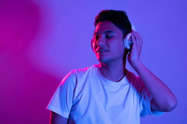 紫色の背景にカラフルな明るいネオンの光の中でポーズを閉じた目でヘッドフォンで音楽を聞いている若いアジアの男の肖像画 — ストック写真