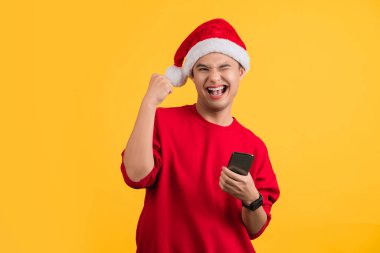 Kırmızı elbiseli ve Noel şapkalı bir adam cep telefonu ve boş ekran çalışma alanı kullanıyor. Sarı arka plan stüdyosunda izole edilmiş jest yapıyor..