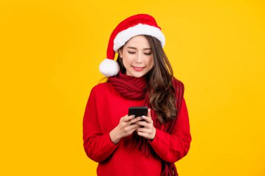Sarı arka planda cep telefonuyla izole edilmiş, noel şapkası takan mutlu genç bir kadının portresi. Mutlu yeni yıl kutlamaları, mutlu tatiller.