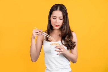 Happy emotion of asian young beautiful woman eat instant noodles cup, isolated over yellow background.