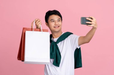 Yakışıklı Asyalı adam akıllı telefonuyla alışveriş çantasıyla fotoğraf çekiyor. Stüdyoda pembe arka planda izole.