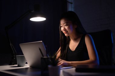 Geceleri masasında dizüstü bilgisayarla çalışan gülümseyen Asyalı kadın.