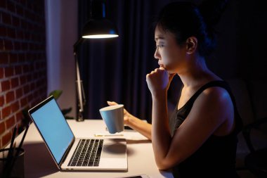 Asyalı kadın çok çalışıyor ve kahve içiyor. Masasında oturmuş karanlıkta dizüstü bilgisayarına bakıyor. Ciddi ciddi düşünüyor..