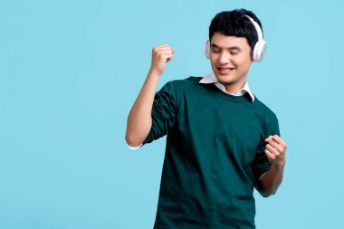 Smiling young handsome asian man listening to the music on cellphone using earbuds, He is dancing isolated in blue background.