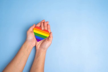 Kadınların elleri açık mavi arka planda gökkuşağının renginde kalbi tutar. LGBT konsepti, Gurur ayı, kopyalama alanı.