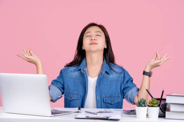 Genç Asyalı iş kadını günlük gömlekler giyer beyaz ofis masasında bilgisayarlı ve pastel pembe arka plan stüdyosunda çalışır. Yogada el ele tutuşuyor. Rahat ol. Meditasyon yap..